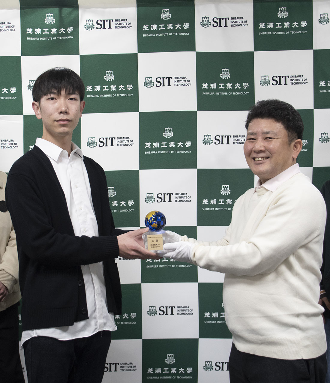 芝浦工業大学のロゴが並んだ背景の前で、トロフィーを持つ若い男性と笑顔の男性が並んで立っています。若い男性は黒いカーディガンと白いシャツを着用し、トロフィーを持っています。右の男性は白い服を着ており、白い手袋をつけてトロフィーを支えています。