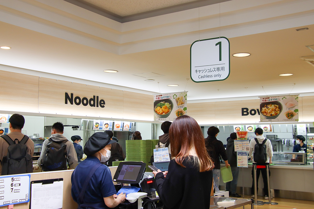 『Noodle』『Bowl』と書かれたカウンターが並ぶ大学の食堂で、スマートフォンを使って会計をする学生と、調理場で働くスタッフの姿