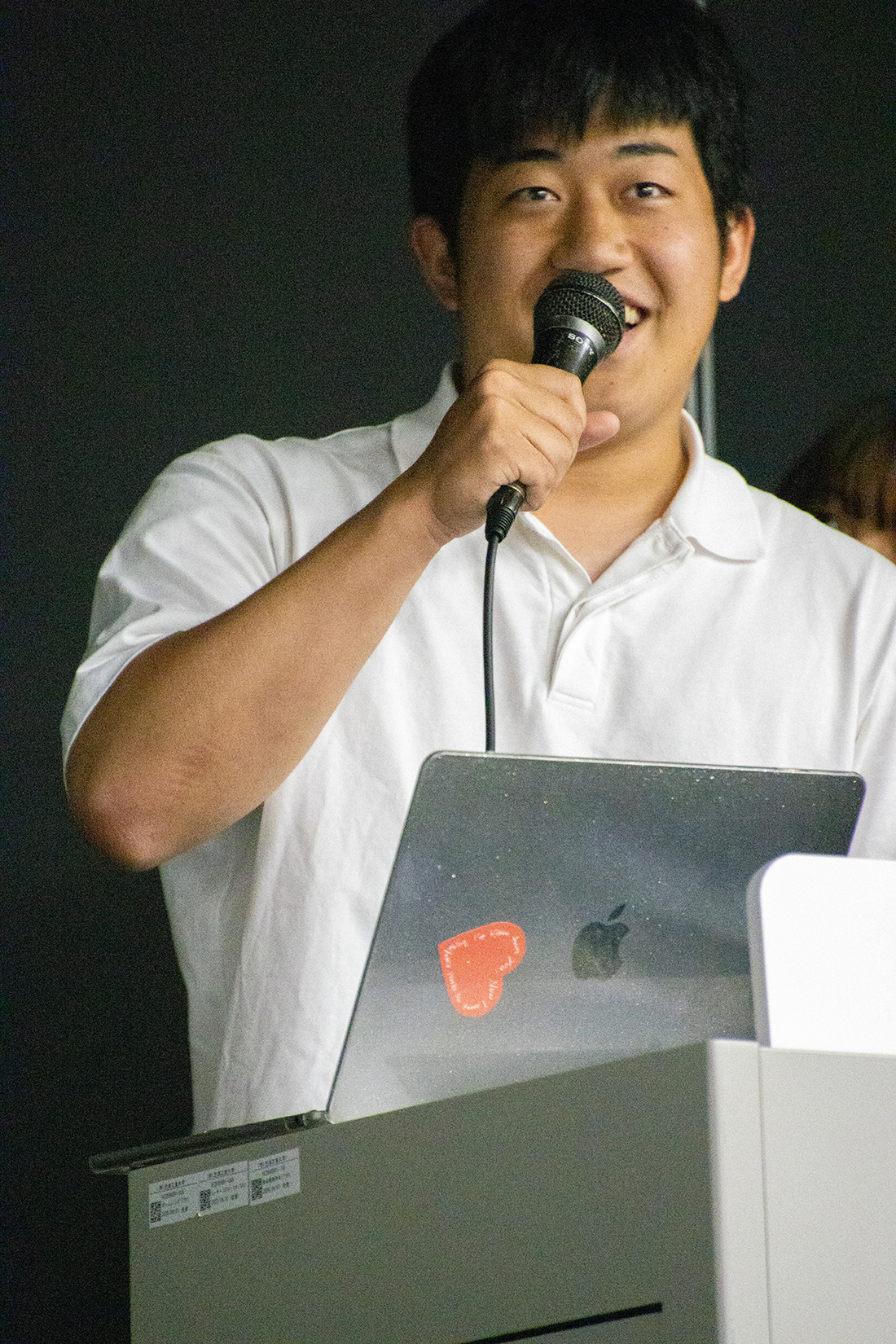 白シャツの人物がマイクを持ち、演台で話している様子