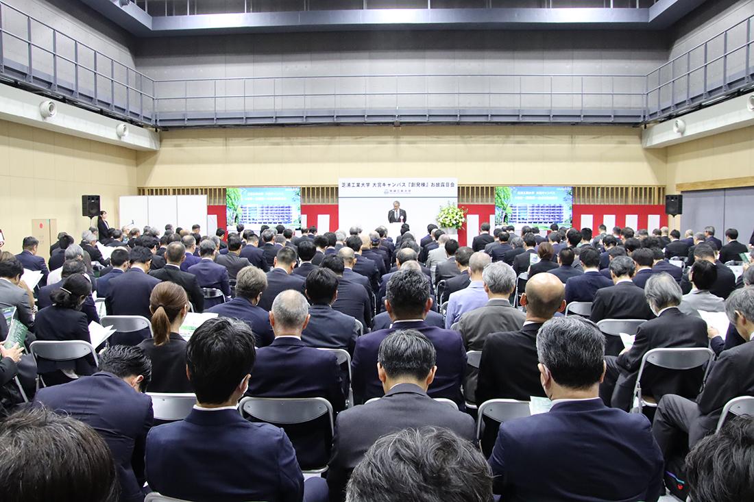 大きな会場で多数が着席し、壇上のあいさつを聞いている 