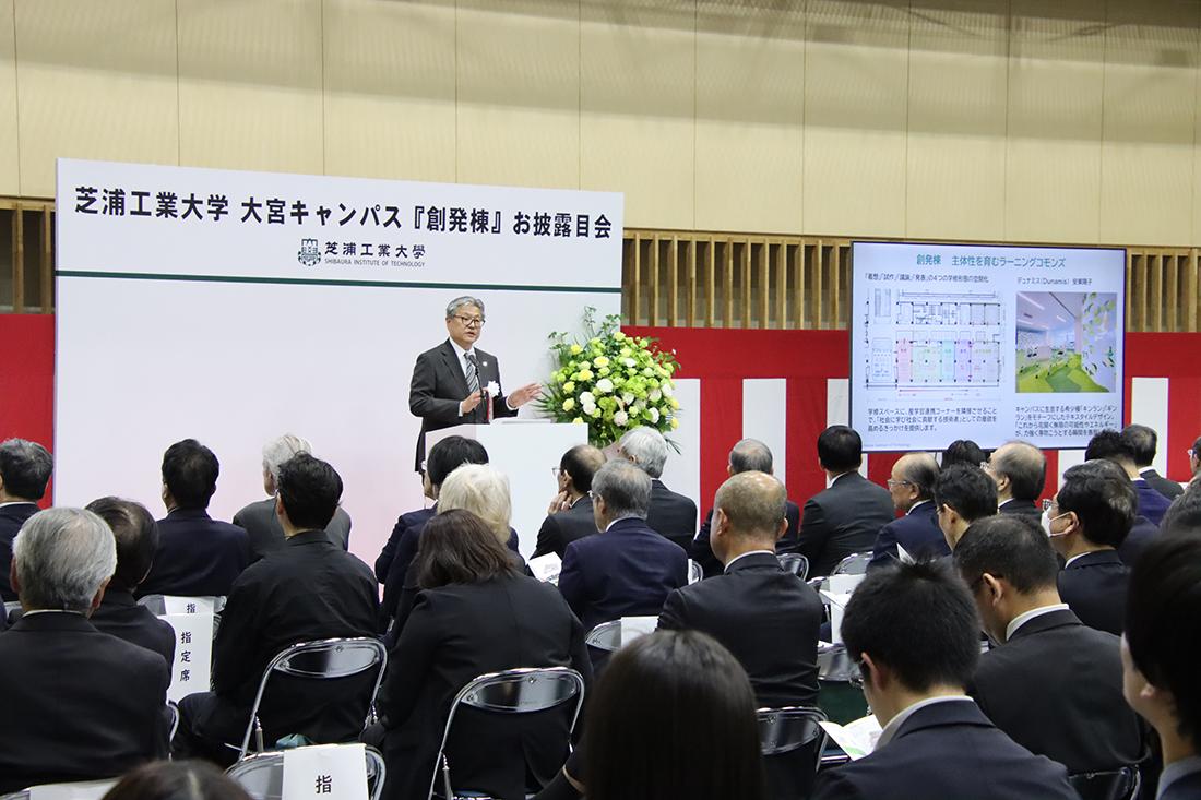 大きな会場で多数が着席し、壇上のあいさつを聞いている 