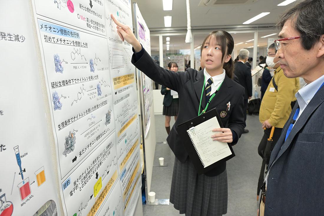 高校生が化学研究のポスターを示し、来場者に解説している