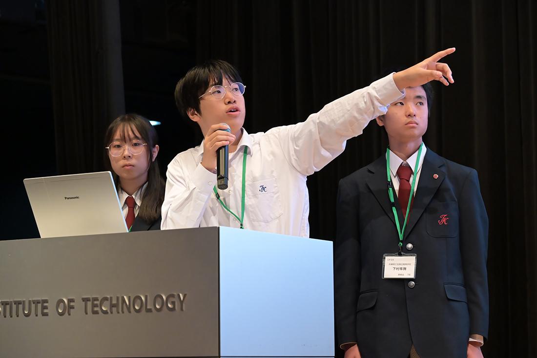 演台で発表者がマイクを持ち、前方を指さして説明している