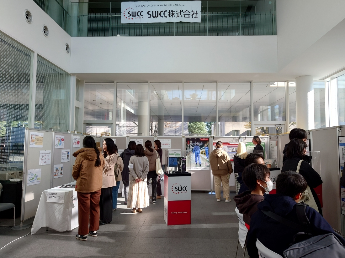展示会場の室内で、複数の人々が様々なブースや展示物を見ている様子。展示パネルに貼られたポスターやテーブルに置かれた資料が見える。