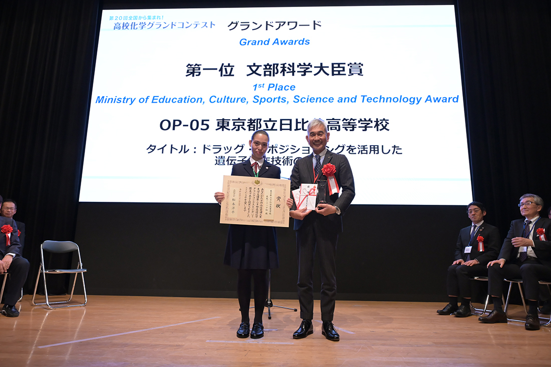 高校化学グランドコンテスト会場の壇上で表彰される受賞者と表彰した学長、後方のディスプレイに映写された表彰内容