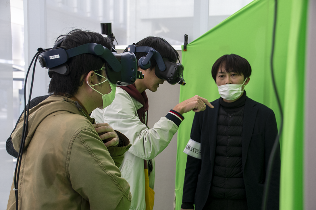 ヘッドマウントディスプレイを装着した2名の人が、緑色の合成用の幕に向かって立っている様子