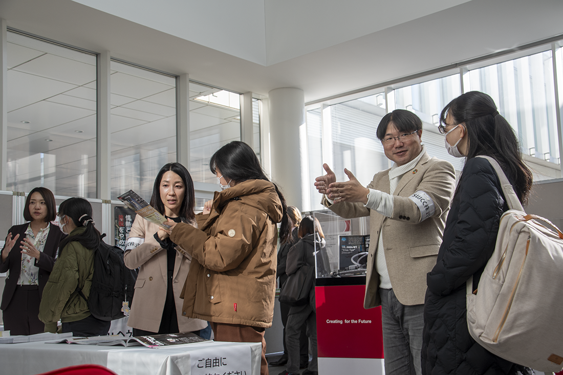 室内で行われている展示イベントの様子。複数の人が資料を手に取り、展示物を見学している。背景にはガラス窓からの自然光が差し込み、明るい雰囲気。