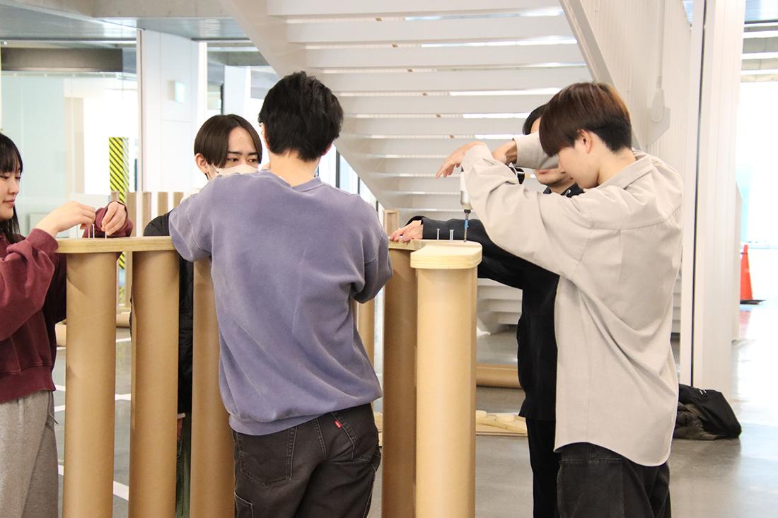 階段横の共有スペースで、複数人が木製構造物のねじ留めをしている