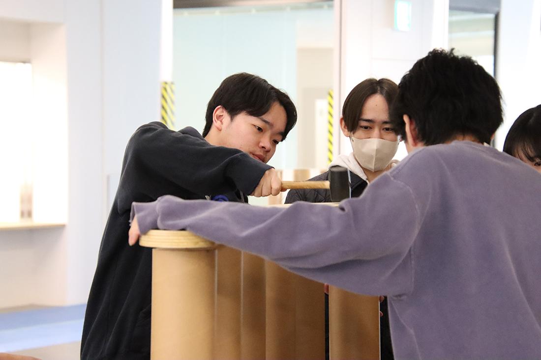 人物が木製の円形天板を押さえ、周囲の人と組み立てをしている