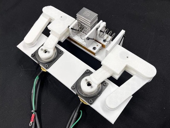 白いフレーム上に、2つの把持機構とばね付き部品が並ぶ実験用装置
